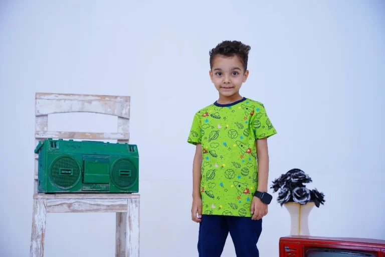 A child stands in front of a white chair with a green vintage boombox, wearing a green space-themed T-shirt, blue pants, and a black smartwatch. A table with black and white flowers and a red retro TV is to the right, against a light blue background.