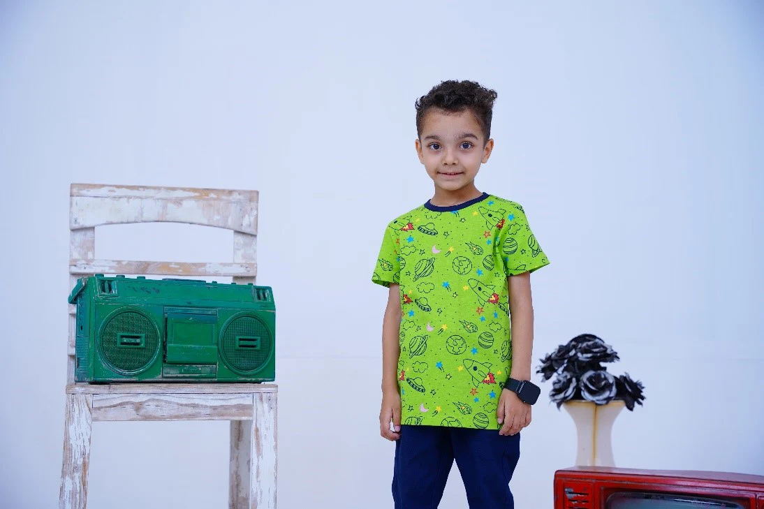 A child stands in front of a white chair with a green vintage boombox, wearing a green space-themed T-shirt, blue pants, and a black smartwatch. A table with black and white flowers and a red retro TV is to the right, against a light blue background.